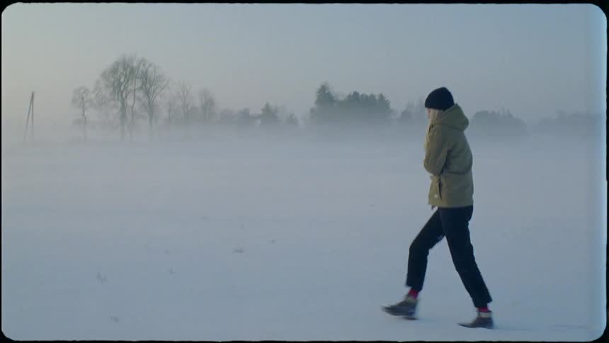 A woman holds herself to keep warm while walking through a snowy field