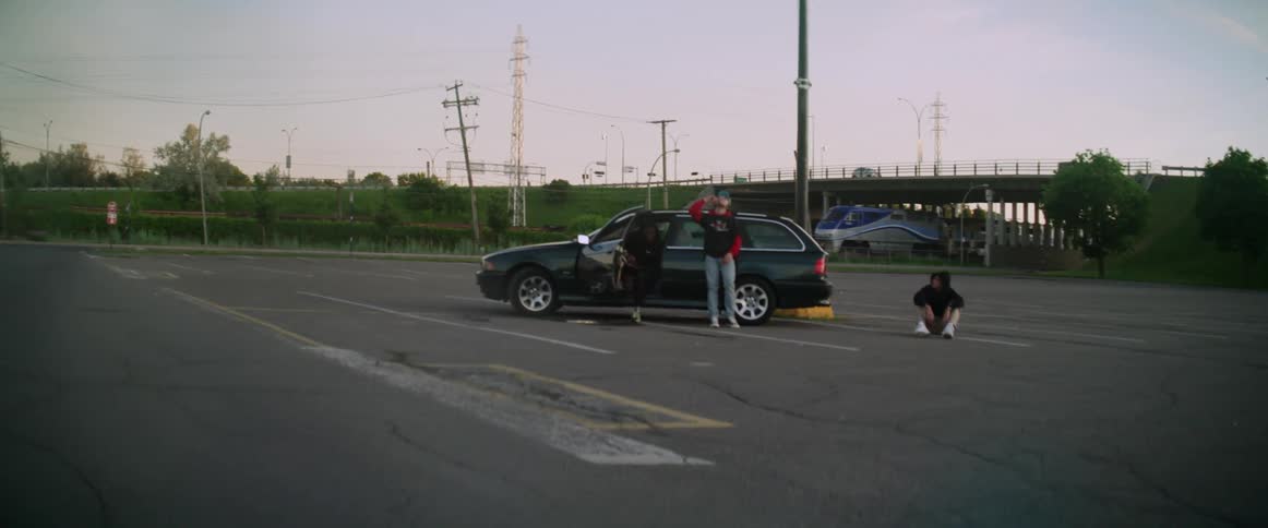 A teenage boy sitting on the ground in a parking lot while his friend leans on a car and another sits in the drivers seat gets up and flips his friends hat off and jokingly pushes him
