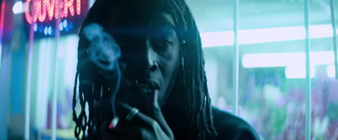 A black man with dread locks is standing outside of a store smoking a cigarette
