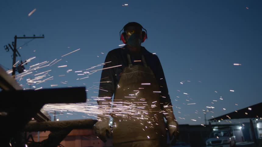 A man is standing wearing a helmet and earmuffs as he watches someone working and sparks fly