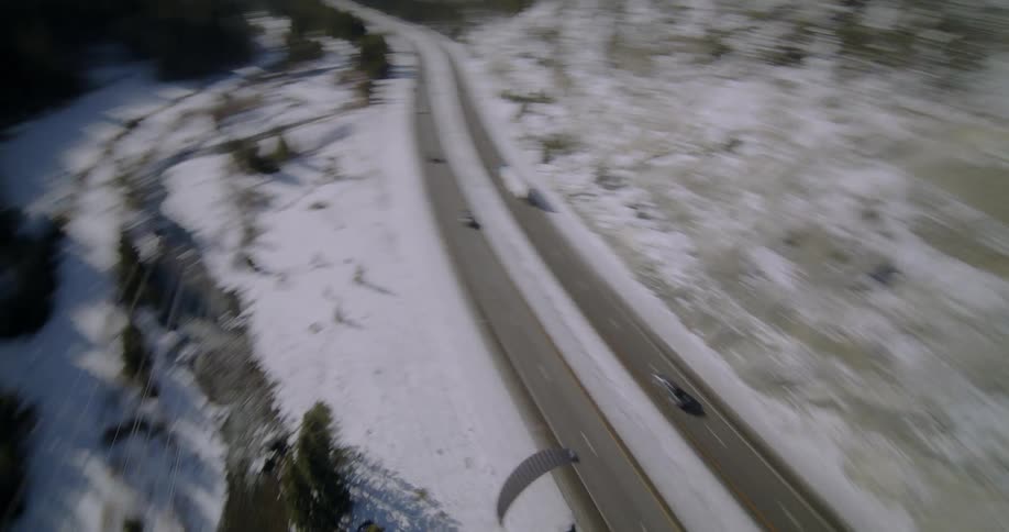 A paraglider is sailing through the air over a highway surrounded by forested mountains as traffic moves below