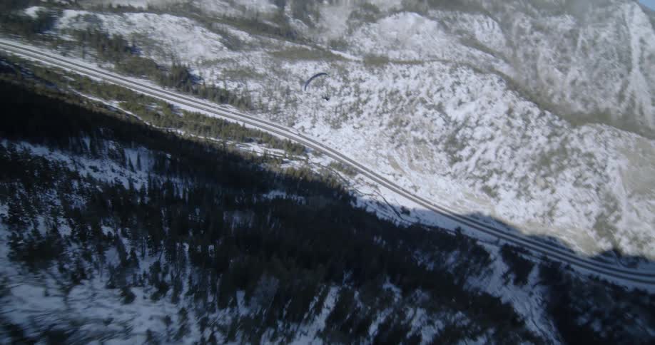 A paraglider is sailing through the air along a forested mountain slope and there is traffic on the highway below
