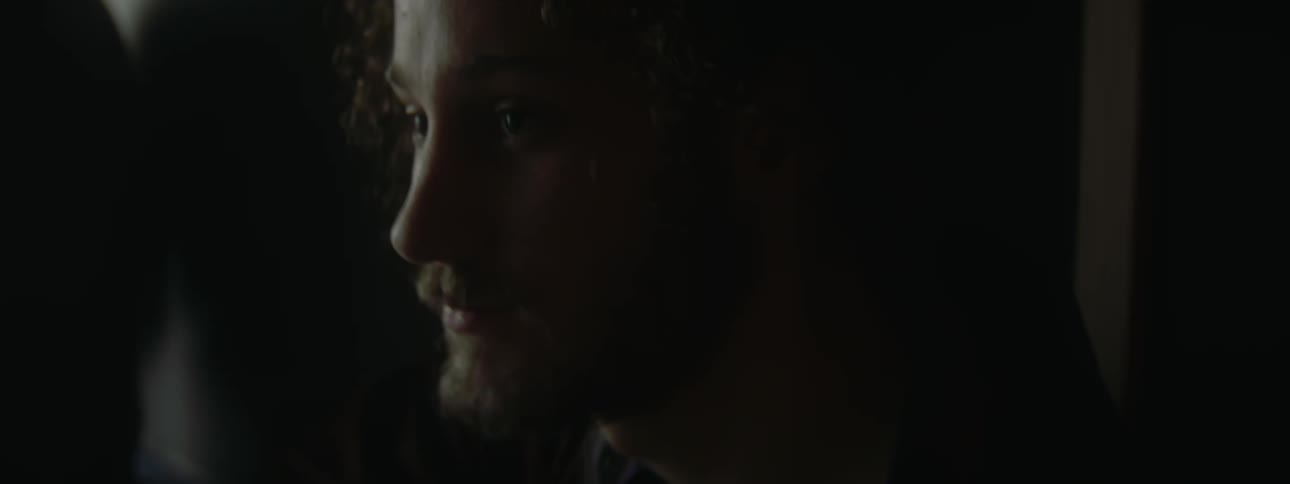 A man with a moustache and beard is sitting in a dark place and there is a light flickering near him