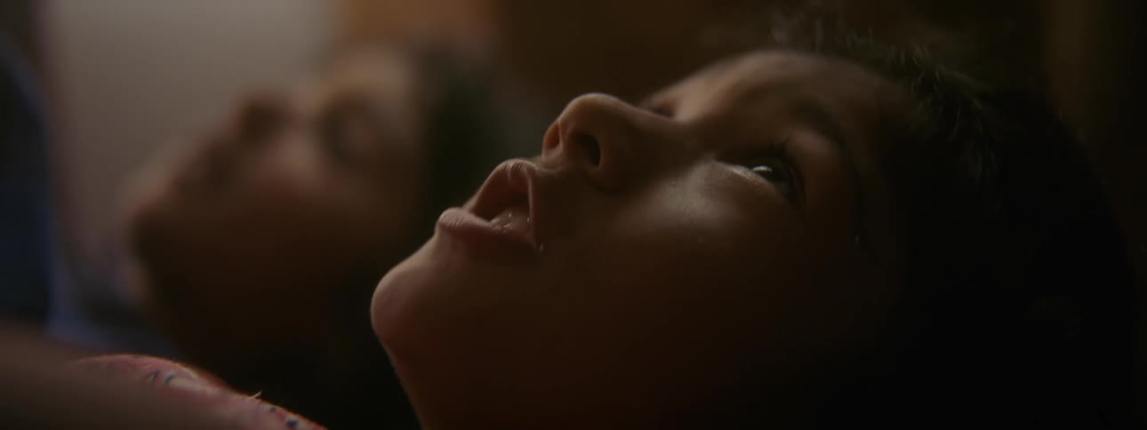 A young girl lies in bed next to her brother and reaches her arms up as she talks to another person