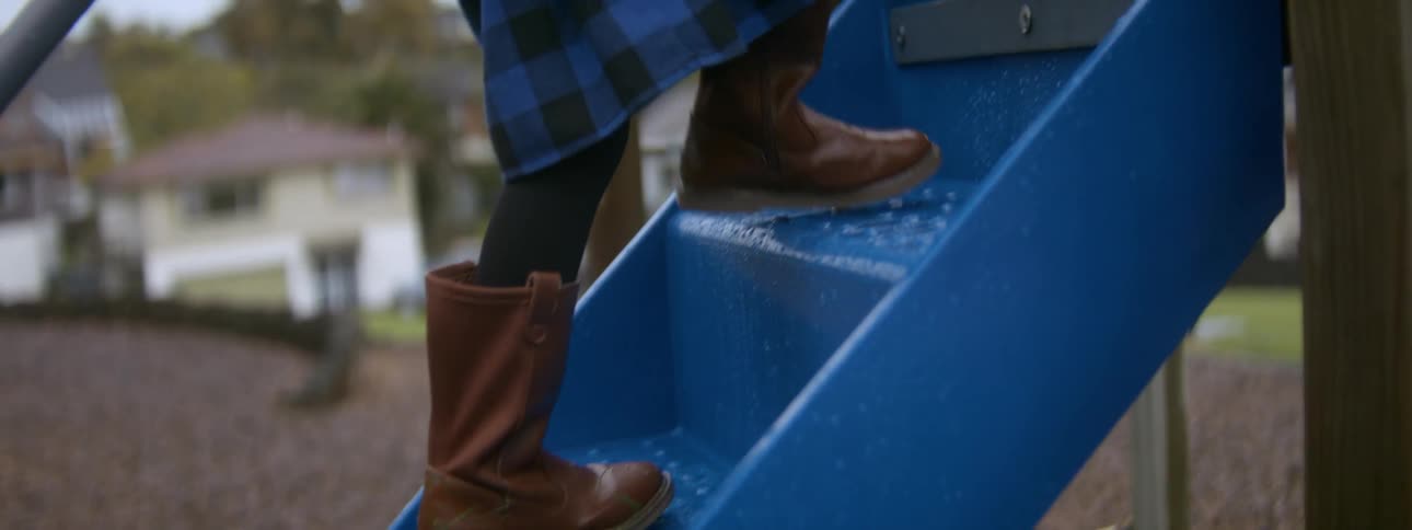 A little girl wearing her school uniform and boots climbs up the steps of playground equipment