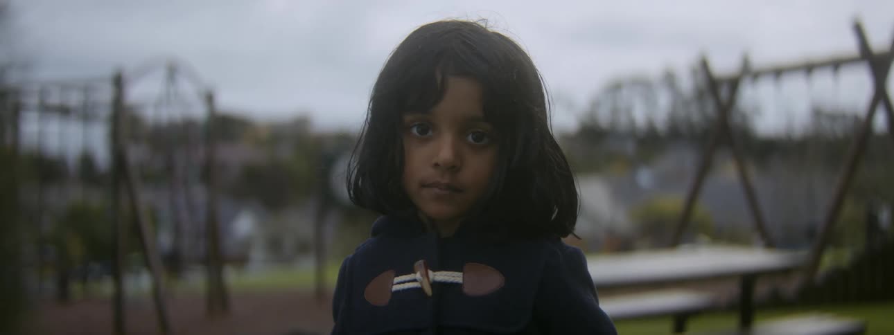 Girl with straight dark wavy hair and navy blue pullover looks at camera in playground