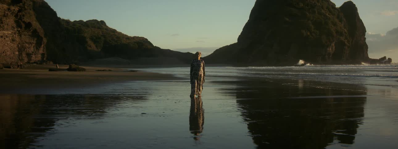 Elderly woman with short hair and long dress walks on wet sand by the sea at sunset