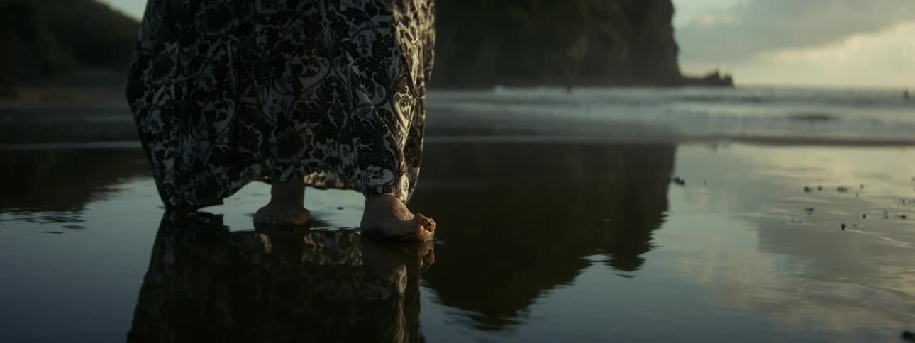 A woman wearing a long print dress walking barefoot on the wet beach sand