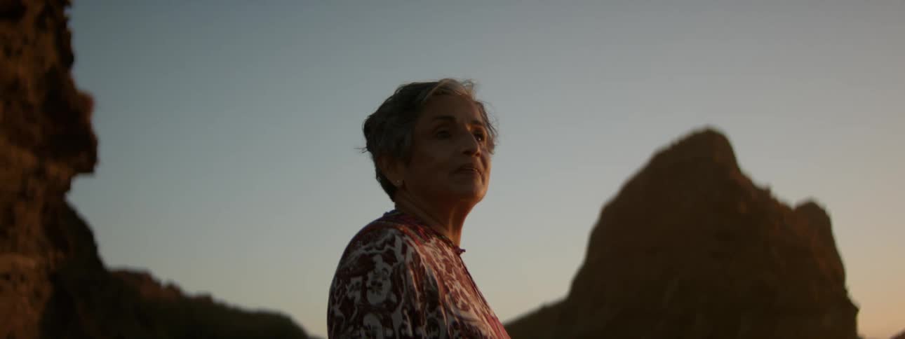 An elderly woman is standing near a rock formation in the mountains