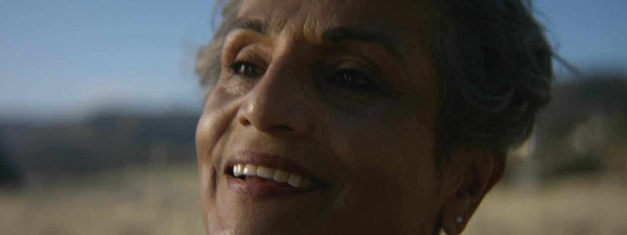 Older woman standing in field smiles as wind blows her hair