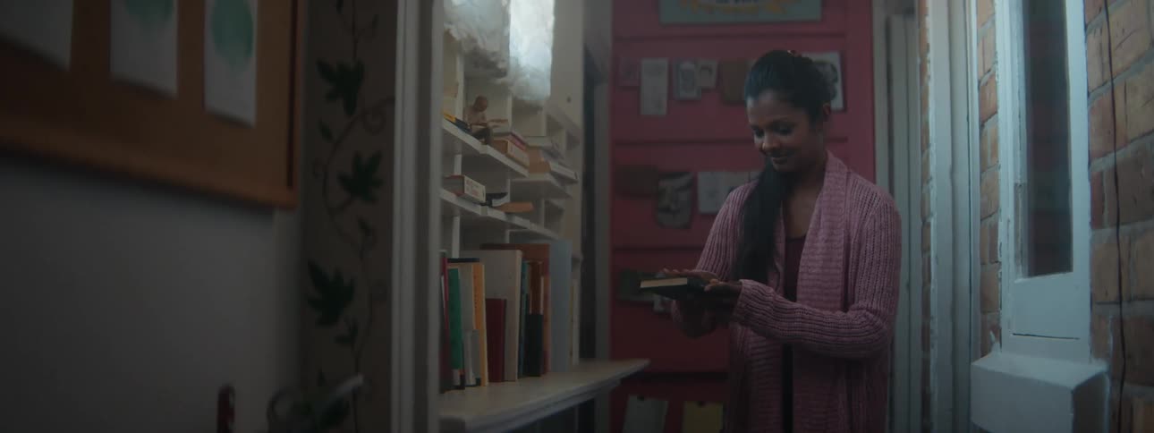 A woman is standing in a hallway looking at books on a shelf as she takes one and wipes the cover with her hand