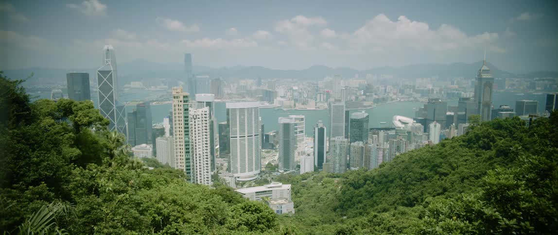 A big city with tall buildings and skyscrapers is seen over a hilltop and there are mountains in the distance under a cloudy sky