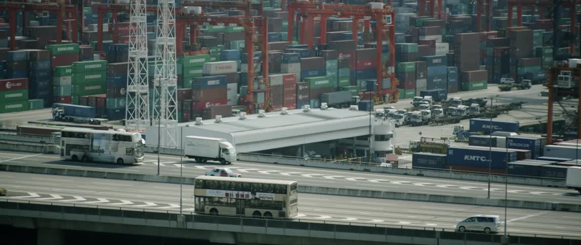 Traffic travels on a highway past a shipping yard full of stacked containers and cranes