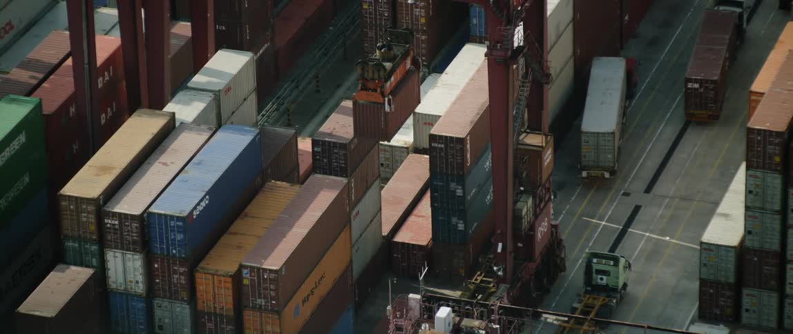 Semi trucks move between stacks of shipping containers as loader slowly lifts one