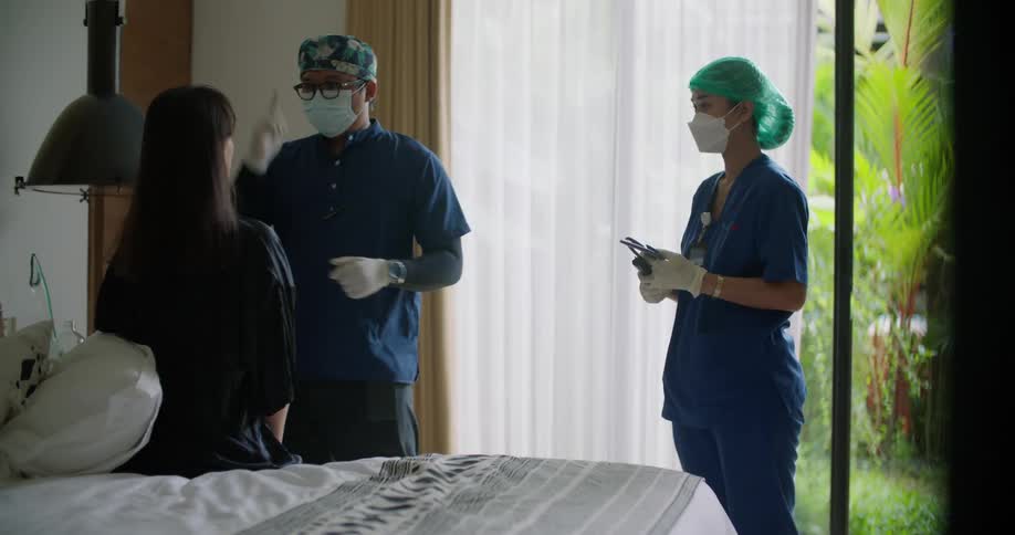 A woman sits on a bed at home as doctor uses a light to look in her mouth and then hands it to a nurse and feels the womans throat