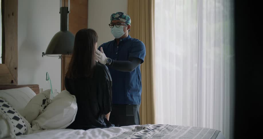 A doctor making a house call feels a womans neck as she sits on a bed