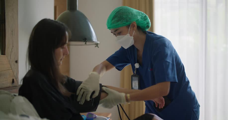 A woman is sitting up in a hospital bed and a nurse is checking her blood pressure