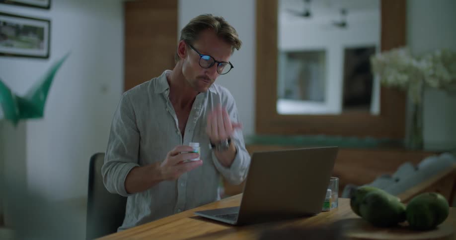 A man takes medication while sitting at his desk behind a laptop