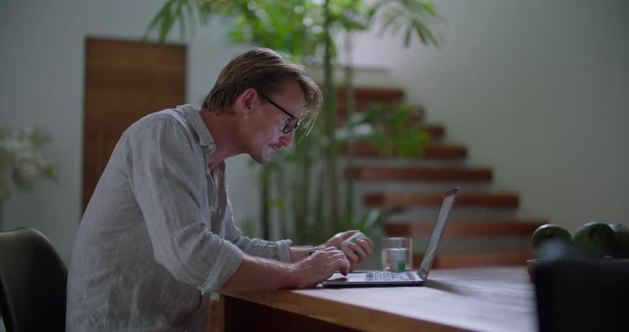 A man using a laptop at a table in a house stops to examine the label of a medicine bottle