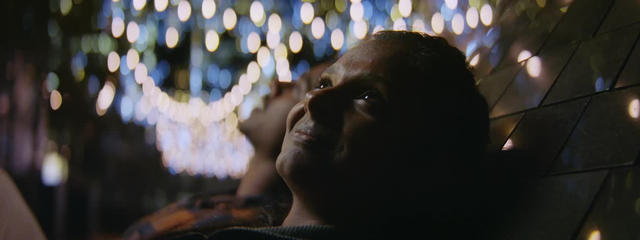 A woman and a man are leaning back sitting in a ride at an amusement park and there are lights in the distance