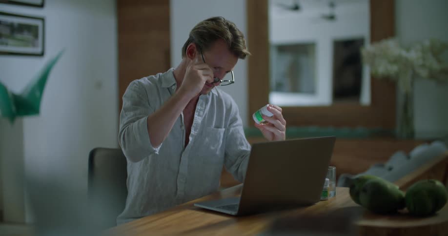 A man is sitting at a desk in front of a laptop computer and he is looking at the label on a medicine bottle