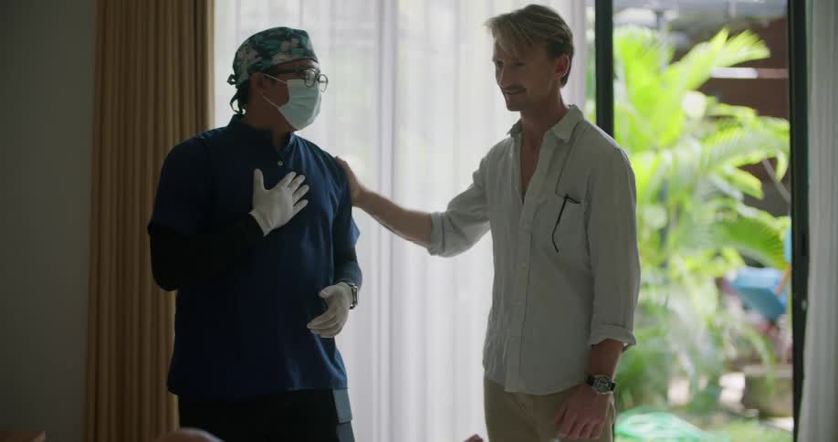 A man is standing in a hospital room talking to a doctor wearing rubber gloves and they shake hands