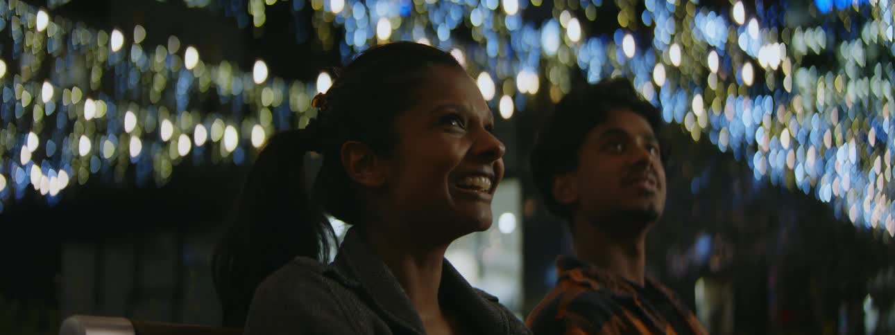 A teenage girl and boy are sitting on a bench talking and there are lights behind them