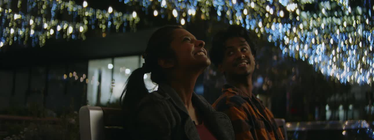 A teenage girl and boy are sitting on a bench talking and there are bright lights on the building behind them