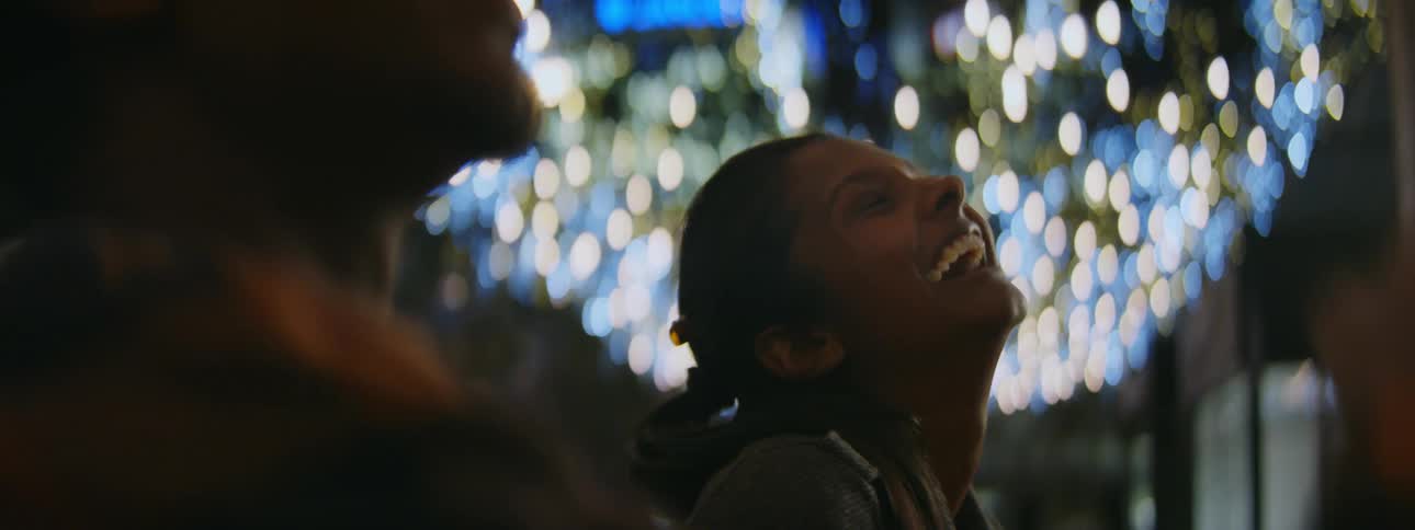 A woman smiles and laughs as she and her male companion stand beneath many bright lights