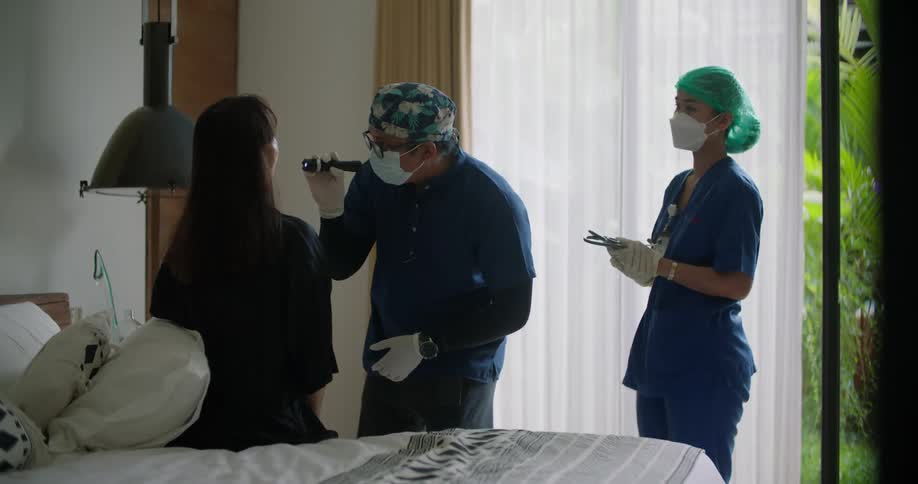 A woman is sitting up in a hospital bed and a nurse hands a doctor a flashlight and he looks inside the patients mouth