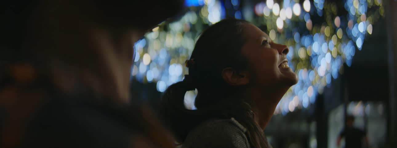 A woman with a man laughs as she looks up at lights outside and a man jogs past them