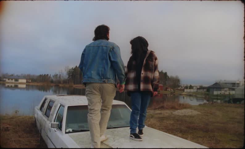A man and woman are next to a lake and they climb onto a car and sit on the roof