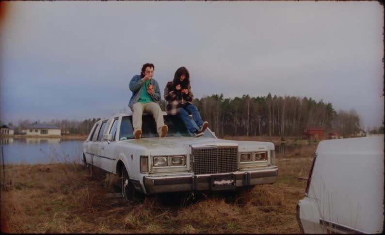 A man and woman eat together while sitting on the roof of a derelict limousine on the shore of a lake