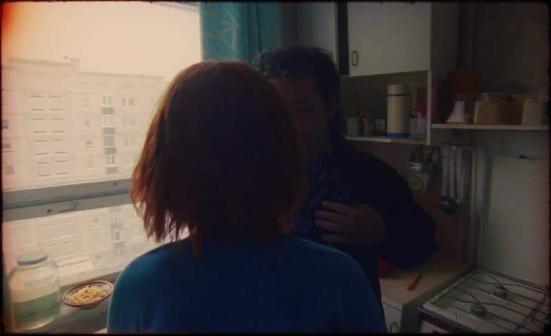 A woman follows her partner into the kitchen and hugs him