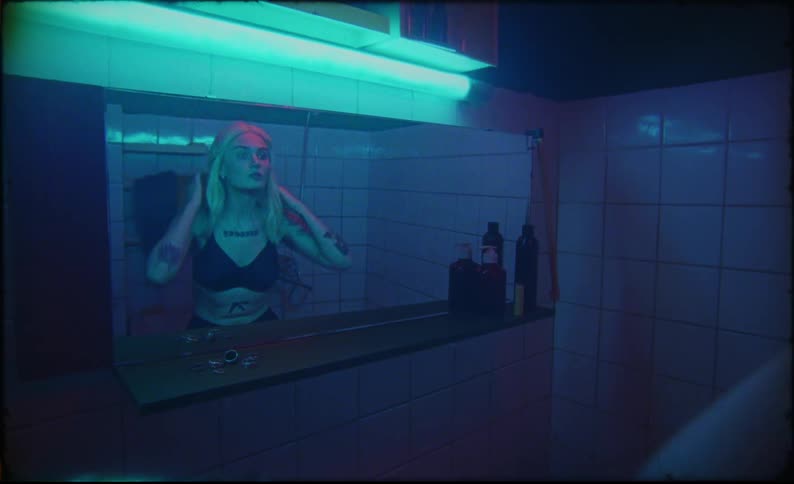 A woman stands in her bathroom as she puts on a long blonde wig in front of a mirror