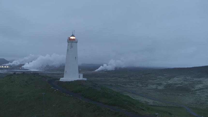 A light moves in a light house as smoke billows from factories in the distance