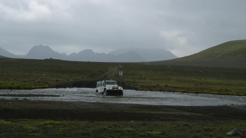 A white jeep slowly drives through a shallow river in a hilly environment