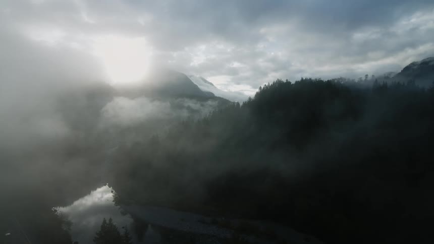 The sun shines through a smoke filled cloudy sky above forested mountains and a lake