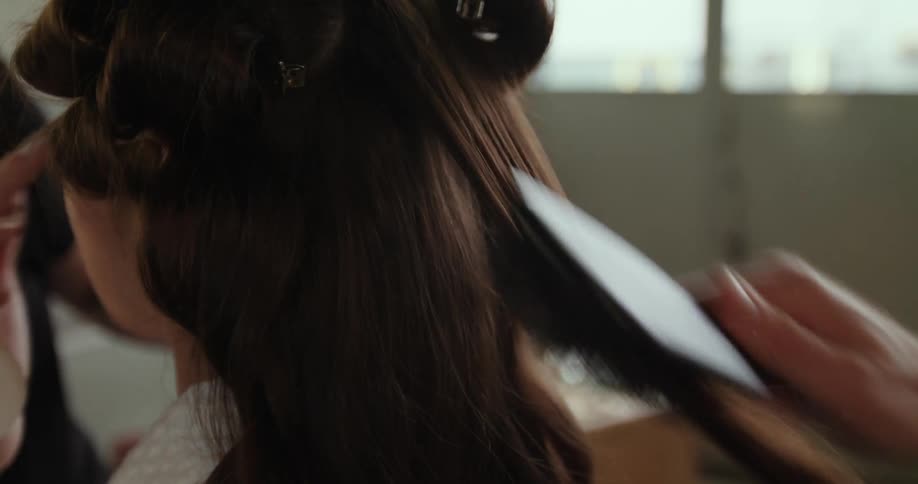 A woman is sitting in a chair and as one person is brushing her long brown hair another person is putting on her makeup