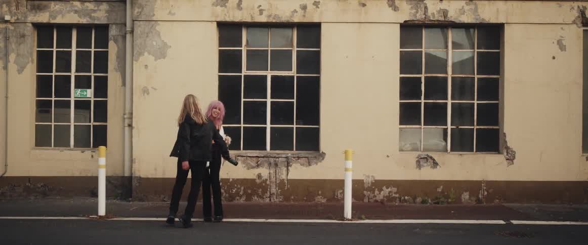 A pink haired woman wearing a white fur coat and a long haired man are walking and playing on a street near buildings each with a bottle in their hands and appear to be drunk