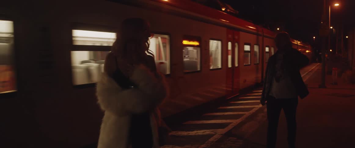 A woman carrying a wine bottle walks beside a subway as it passes and a man follows behind her