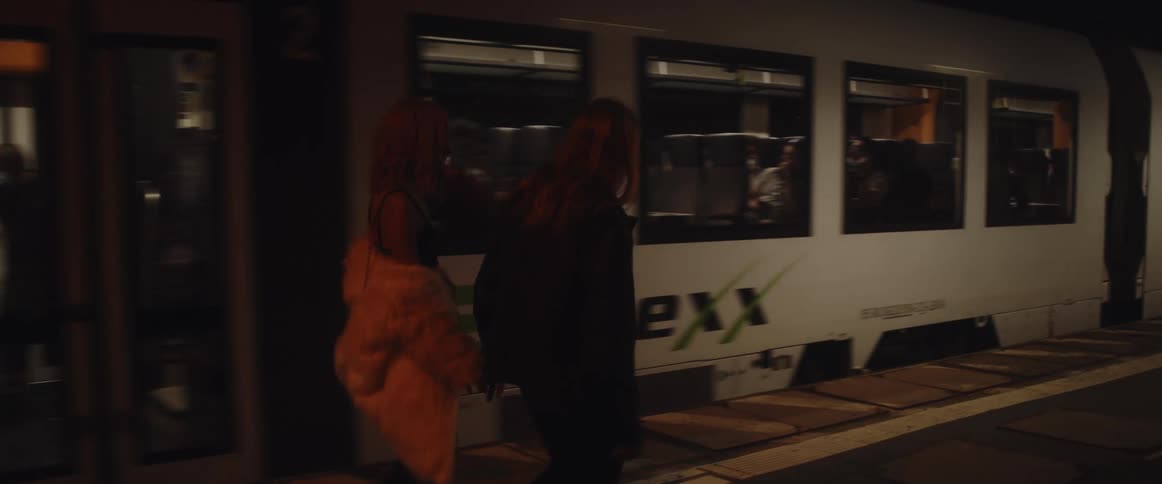 A redheaded woman wearing a white fur coat and a long haired man are walking near a moving subway train
