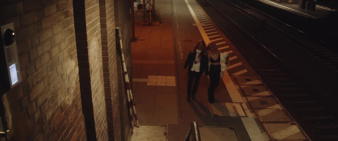 A woman carrying a bottle of wine holds a mans hand and the intoxicated couple stumble down the street