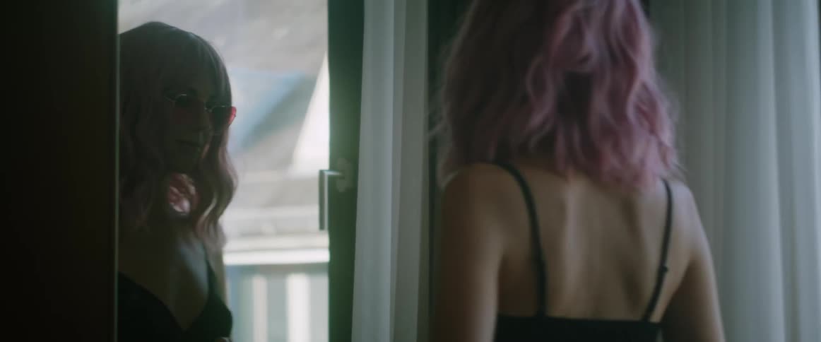 A young woman grins while looking at her reflection in a mirror before turning and walking away