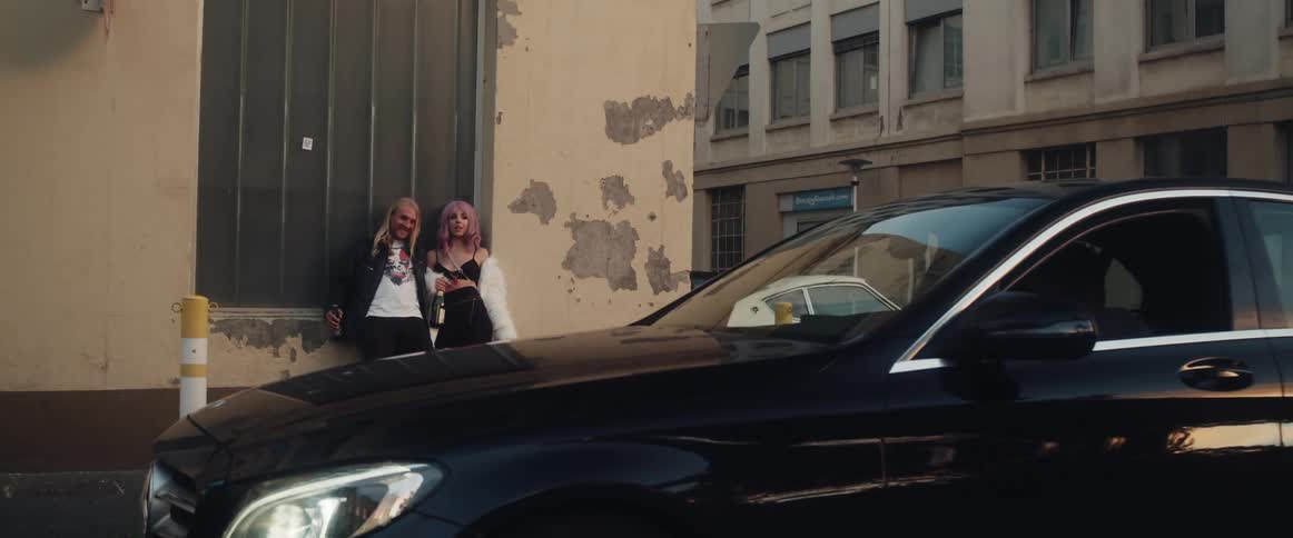 A man and woman holding bottles of booze lean against a building and the woman leans over and kisses him as a car pulls up