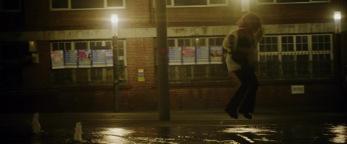 A woman is holding a wine bottle and jumping and dancing in the rain on a street near buildings