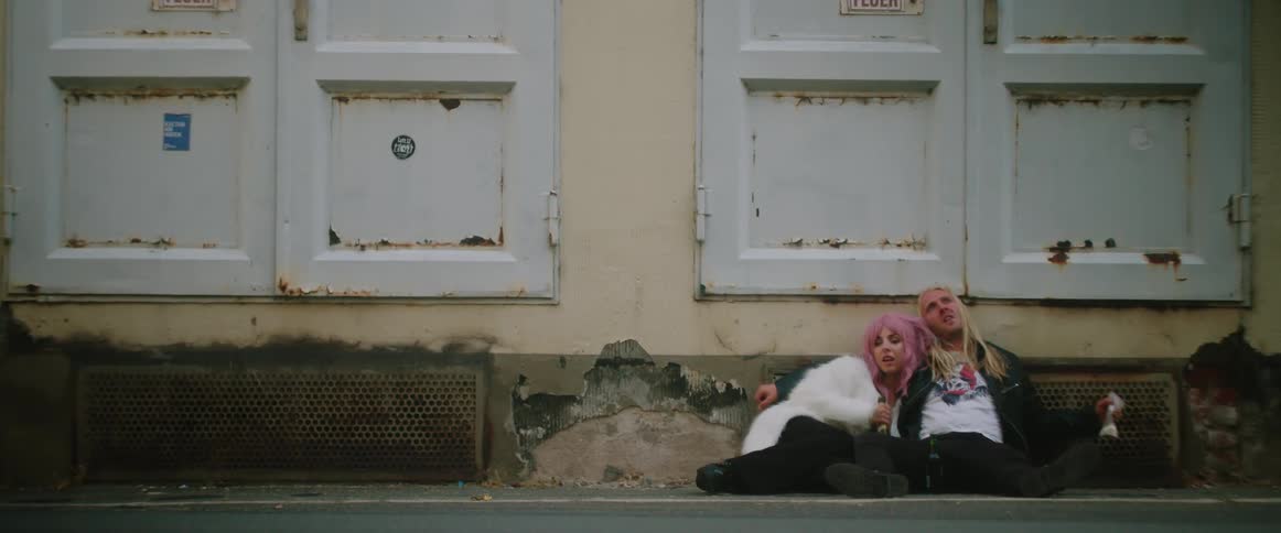 A pink haired woman wearing a white fur coat and a long haired man are sitting outside leaning against the wall of an abandoned building and she is holding a wine bottle and he has a bag of white powder