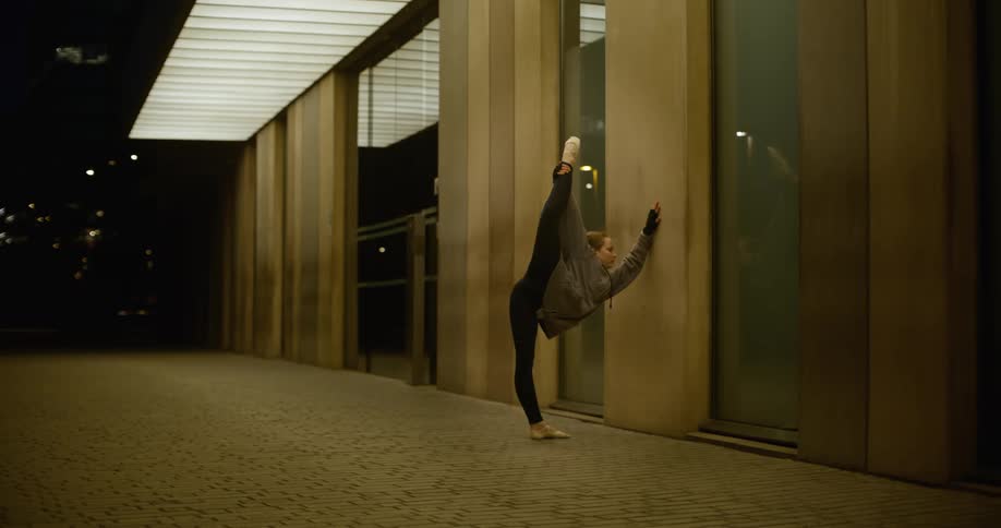 A teenage girl is practicing ballet as she leans her arm on the wall and holds her foot in the air above her head