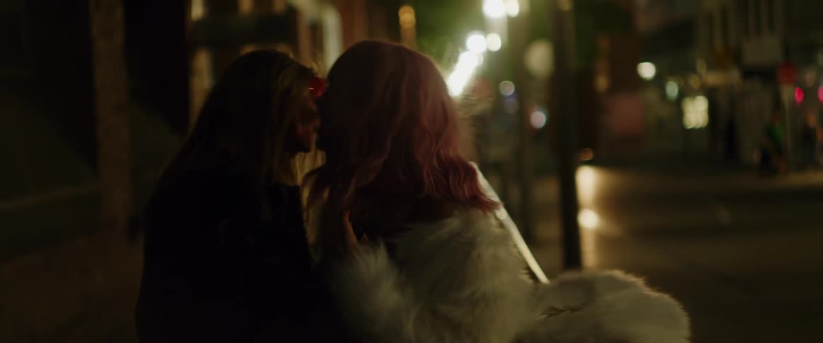 A redheaded woman wearing a white fur coat and carrying a bottle of wine is talking to a man with long blond hair and they walk down the sidewalk