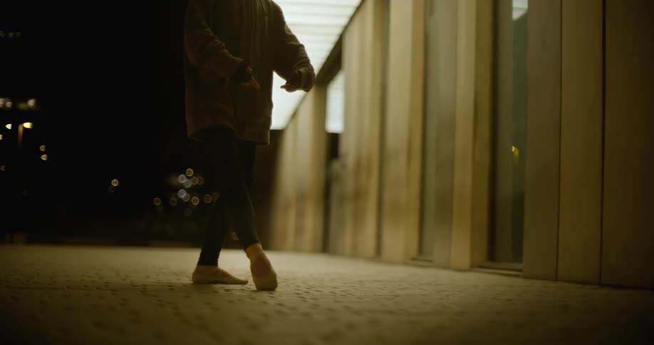 A person practicing ballet dance steps near a lit building at night